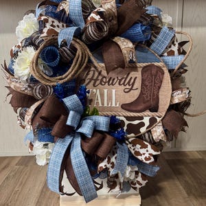 May include: A decorative wreath with a rustic, Western theme. It features brown, blue, and white ribbons, cowhide print accents, and a rope detail. The centerpiece has a brown cowboy boot and the text "Howdy Y'all".