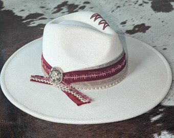 Pode incluir: Um chapéu fedora branco de aba larga, decorado com uma fita de veludo bordô, uma fita bege com lantejoulas rosa e um botão prateado.