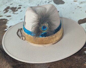 Pode incluir: Um chapéu fedora de feltro bege com uma faixa de veludo marrom e uma fita de veludo azul. Uma pena de pavão com olhos azuis e verdes está presa ao chapéu.