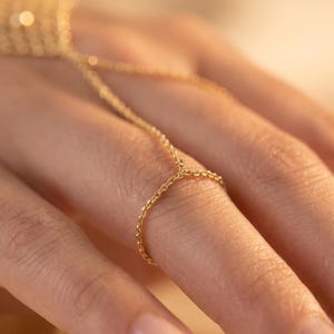 Puede incluir: Una delicada cadena de mano dorada, con un anillo conectado por una fina cadena a un dise&ntilde;o tipo pulsera. La joya est&aacute; elaborada con una cadena sutil y texturizada, creando una est&eacute;tica minimalista y elegante. La c&aacute;lida iluminaci&oacute;n realza el brillo del oro.