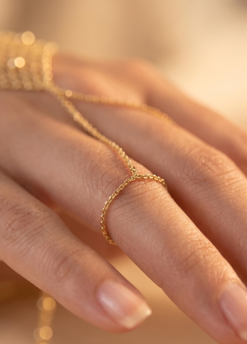 Puede incluir: Una delicada cadena de mano dorada, con un anillo conectado por una fina cadena a un dise&ntilde;o tipo pulsera. La joya est&aacute; elaborada con una cadena sutil y texturizada, creando una est&eacute;tica minimalista y elegante. La c&aacute;lida iluminaci&oacute;n realza el brillo del oro.