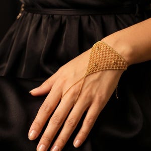 Puede incluir: Un brazalete de cadena de mano dorado. El brazalete presenta un delicado dise&ntilde;o de red con peque&ntilde;os acentos redondos. Una fina cadena se extiende desde el brazalete hasta un anillo para el dedo. El fondo es oscuro.