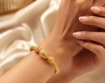 Puede incluir: Una pulsera dorada con una banda texturizada similar a una malla, adornada con tres dijes esféricos. Cada dije muestra un intrincado patrón de filigrana perforada. La pulsera tiene un acabado metálico pulido y un diseño circular.
