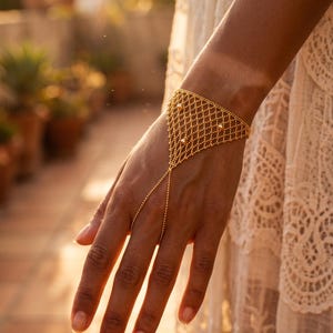 Puede incluir: Una pulsera de cadena de mano dorada. La pulsera presenta un dise&ntilde;o geom&eacute;trico de cuentas interconectadas en la mu&ntilde;eca, con una delicada cadena que se extiende hasta un anillo para el dedo. Peque&ntilde;as cuentas decorativas est&aacute;n intercaladas en el dise&ntilde;o.