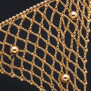 Puede incluir: Primer plano de un accesorio de estilo cota de malla de color dorado. El intrincado dise&ntilde;o presenta un patr&oacute;n en forma de diamante creado por cadenas interconectadas. Varias peque&ntilde;as cuentas redondas de color dorado se intercalan en el dise&ntilde;o, con cuentas esf&eacute;ricas m&aacute;s grandes a intervalos.