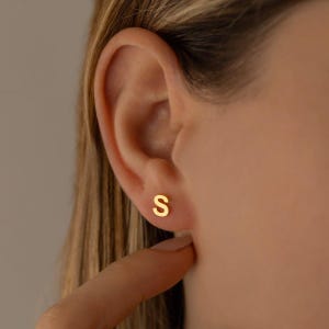 May include: A small gold-toned stud earring shaped like the capital letter S. The earring is positioned on the earlobe, featuring a smooth, polished surface and a simple, clean design that sits flush against the skin for a subtle appearance.