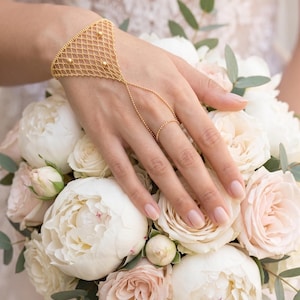 Puede incluir: Una pulsera de cadena de mano dorada con un delicado dise&ntilde;o de red y peque&ntilde;os detalles de cuentas. La pulsera se conecta a un anillo a trav&eacute;s de una fina cadena. La mano descansa sobre un ramo de rosas y peon&iacute;as blancas y rosadas.