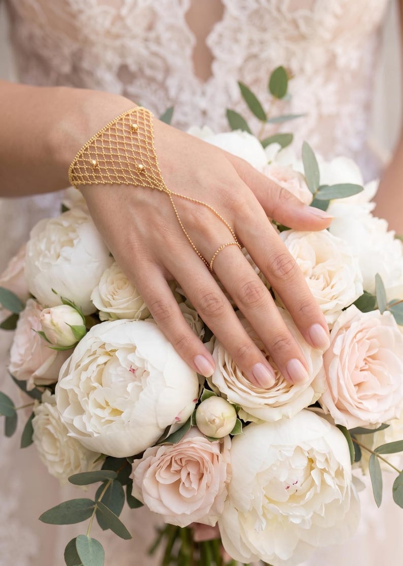 Puede incluir: Una pulsera de cadena de mano dorada con un delicado dise&ntilde;o de red y peque&ntilde;os detalles de cuentas. La pulsera se conecta a un anillo a trav&eacute;s de una fina cadena. La mano descansa sobre un ramo de rosas y peon&iacute;as blancas y rosadas.