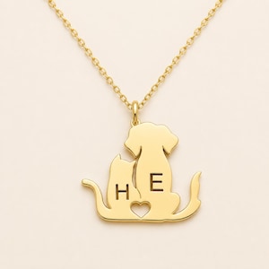 Pode incluir: Um colar dourado com um pingente em forma das silhuetas de um gato e um cão sentados lado a lado. A silhueta do gato está gravada com a letra H, a do cão com a letra E, e um pequeno coração recortado está posicionado no centro.