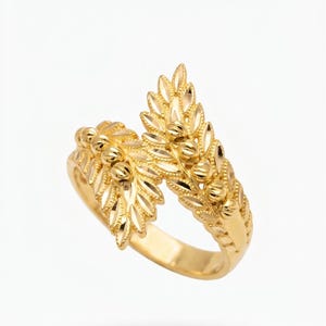Könnte beinhalten: Ein goldfarbener Ring mit einem detaillierten Blattdesign. Der Ring hat ein Band und zwei verzierte blattartige Strukturen, die sich nach oben wölben. Die Blätter sind mit kleinen, perlenartigen Akzenten strukturiert.