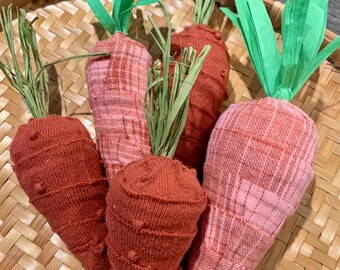 May include: A collection of five handmade carrot decorations. The carrots are made from fabric in shades of orange and pink, with green paper and dried grass for the tops. They are arranged in a woven basket.