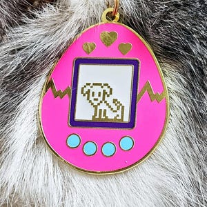 May include: A pink and gold enamel pendant resembling a retro digital pet toy. It features a pixelated dog graphic on a white background, with three gold hearts and three blue circles. A gold-coloured ring is attached at the top.