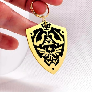 May include: A gold-colored shield-shaped keychain with a black design featuring a triangle and a bird emblem. The keychain has a gold ring for attaching keys. The design is a stylized representation of a shield.