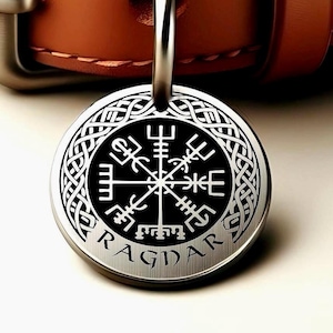May include: A round, silver-colored pendant with a black center featuring a white Viking symbol. The pendant has a braided border and the name "RAGDAR" is engraved around the edge. The pendant is attached to a silver loop and a brown leather strap.