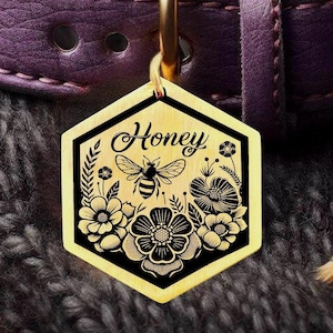 May include: A gold-coloured, hexagonal pet tag with the word "Honey" in script. The tag features a bee and floral design in black. The tag is attached to a purple leather collar.
