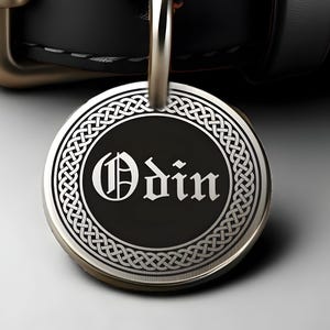 May include: A silver-colored, circular pendant with the name "Odin" in a stylized font. The pendant has a black center and a Celtic knot border. A silver loop is attached at the top.
