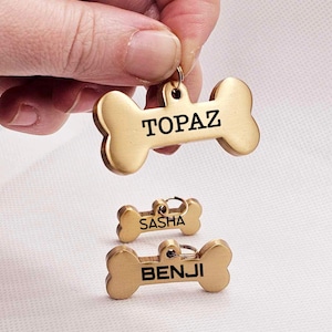 May include: Gold-coloured bone-shaped pet tags with names engraved in black. The top tag reads "TOPAZ", held by a hand. Below are tags that read "SASHA" and "BENJI". The tags have a small loop for attaching to a collar.