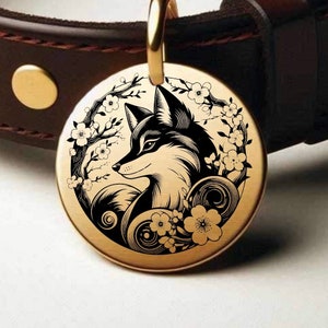 May include: A gold-colored, round pet tag with a black and white fox illustration surrounded by floral branches. The tag is attached to a brown leather collar with gold hardware.