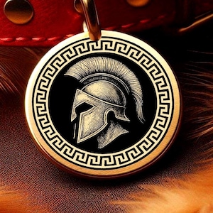 Könnte beinhalten: Eine runde, goldfarbene Haustiermarke mit einem schwarzen Zentrum, das eine detaillierte Illustration eines spartanischen Helms zeigt. Die Marke hat einen griechischen Schlüsselrand und ist an einem roten Lederhalsband befestigt.