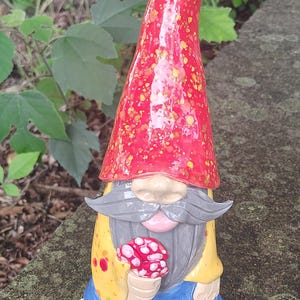 Może przedstawiać: Ceramiczna figurka krasnala ogrodowego z wysoką, spiczastą czerwoną czapką w ż&oacute;łte kropki, długimi szarymi wąsami i brodą, ż&oacute;łtą koszulą w czerwone plamy, niebieskimi spodniami i bosymi stopami, trzymająca w dłoniach małego czerwono-białego grzyba.