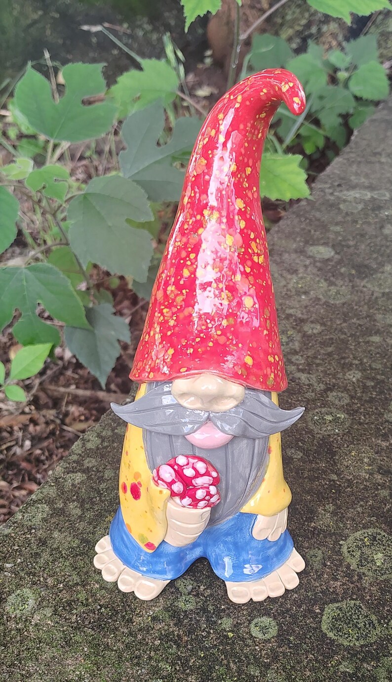 Może przedstawiać: Ceramiczna figurka krasnala ogrodowego z wysoką, spiczastą czerwoną czapką w ż&oacute;łte kropki, długimi szarymi wąsami i brodą, ż&oacute;łtą koszulą w czerwone plamy, niebieskimi spodniami i bosymi stopami, trzymająca w dłoniach małego czerwono-białego grzyba.