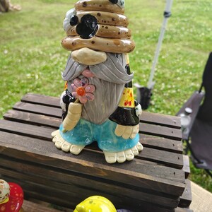Może przedstawiać: Ceramiczna figurka gnoma w beżowym, nakrapianym kapeluszu w kształcie ula z czarnymi akcentami. Gnom ma długą szarą brodę, niebieskie spodnie i trzyma mały r&oacute;żowy kwiatek. Posiada kremowe dłonie i stopy z widocznymi palcami u n&oacute;g.