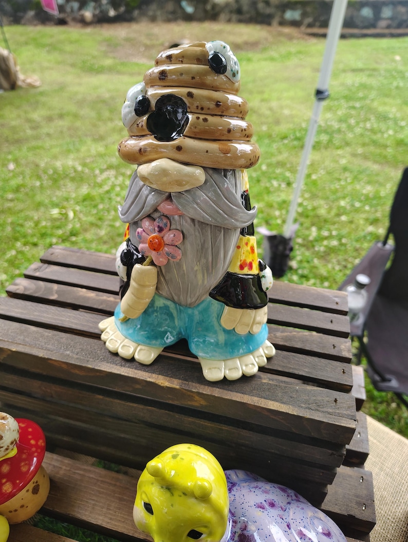 Może przedstawiać: Ceramiczna figurka gnoma w beżowym, nakrapianym kapeluszu w kształcie ula z czarnymi akcentami. Gnom ma długą szarą brodę, niebieskie spodnie i trzyma mały r&oacute;żowy kwiatek. Posiada kremowe dłonie i stopy z widocznymi palcami u n&oacute;g.