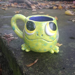 May include: A whimsical ceramic mug shaped like a frog. The mug is primarily yellow with green speckles and a blue interior. It features large, expressive eyes and small feet, creating a playful design. The handle is curved and matches the mug's color scheme.