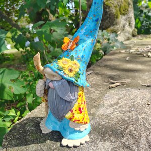 Może przedstawiać: Ceramiczna figurka gnoma z wysokim, nakrapianym niebieskim spiczastym kapeluszem ozdobionym małym pomarańczowym motylem i ż&oacute;łtymi słonecznikami. Gnom ma długą szarą brodę, jasnoniebieską tunikę, ż&oacute;łty wzorzysty płaszcz i trzyma w dłoni drewnianą laskę.