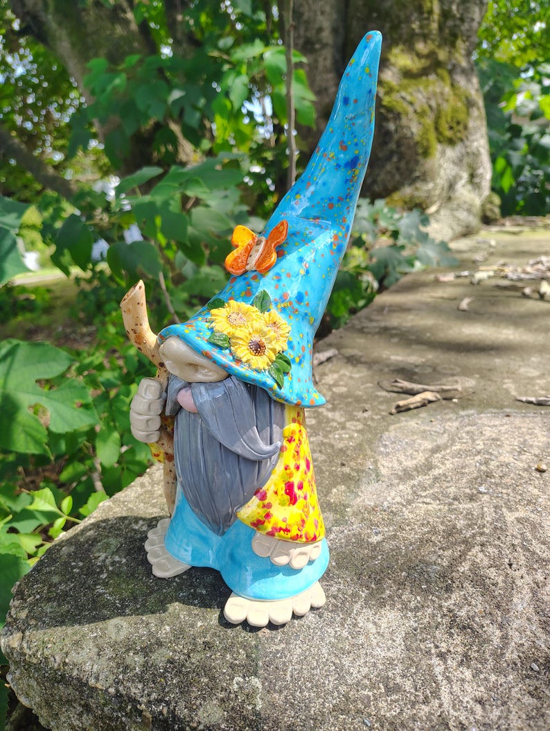 Może przedstawiać: Ceramiczna figurka gnoma z wysokim, nakrapianym niebieskim spiczastym kapeluszem ozdobionym małym pomarańczowym motylem i ż&oacute;łtymi słonecznikami. Gnom ma długą szarą brodę, jasnoniebieską tunikę, ż&oacute;łty wzorzysty płaszcz i trzyma w dłoni drewnianą laskę.