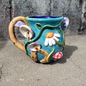 Könnte beinhalten: Eine tealblaue Keramiktasse mit einem fantasievollen Design mit Pilzen, Ranken und einer weißen Margerite. Der Henkel ist wie eine Ranken mit braunen Flecken geformt.