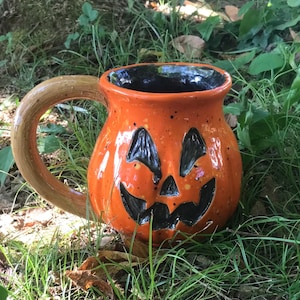 Puede incluir: Taza de cerámica naranja con forma de calabaza con una cara negra y un asa marrón.