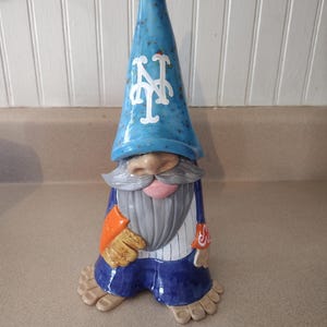 Może przedstawiać: Ceramiczna figurka gnoma w niebieskiej spiczastej czapce z białym logo NY, szarymi wąsami i brodą oraz w białej koszuli w paski i niebieskich spodniach. Gnom ma beżowy nos, r&oacute;żowe usta i beżowe stopy z widocznymi palcami, trzymając beżową rękawicę.