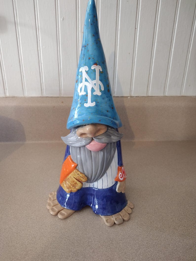 Może przedstawiać: Ceramiczna figurka gnoma w niebieskiej spiczastej czapce z białym logo NY, szarymi wąsami i brodą oraz w białej koszuli w paski i niebieskich spodniach. Gnom ma beżowy nos, r&oacute;żowe usta i beżowe stopy z widocznymi palcami, trzymając beżową rękawicę.