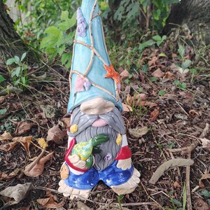 Może przedstawiać: Ceramiczna figurka krasnala ogrodowego z wysoką, teksturowaną jasnoniebieską czapką ozdobioną małymi muszelkami i rozgwiazdą. Krasnal ma długą szarą brodę, niebieskie spodnie i trzyma w dłoniach małego zielono-ż&oacute;łtego ż&oacute;łwia morskiego.