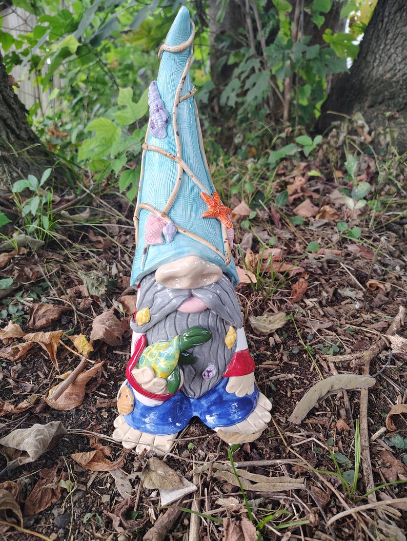 Może przedstawiać: Ceramiczna figurka krasnala ogrodowego z wysoką, teksturowaną jasnoniebieską czapką ozdobioną małymi muszelkami i rozgwiazdą. Krasnal ma długą szarą brodę, niebieskie spodnie i trzyma w dłoniach małego zielono-ż&oacute;łtego ż&oacute;łwia morskiego.