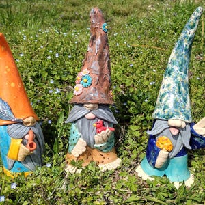 May include: Three ceramic garden gnomes with tall, pointed hats. One gnome has an orange hat and a feather, another has a brown hat with flowers, and the third has a blue and green hat. They are in a grassy area.