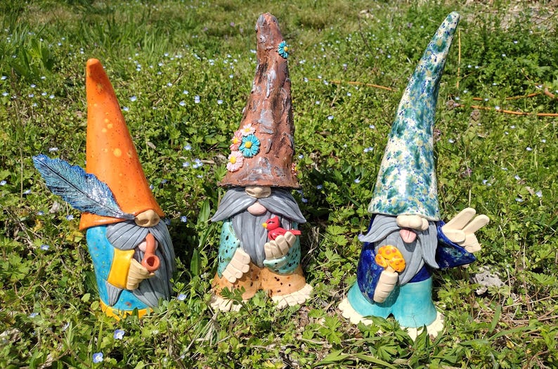 Może przedstawiać: Trzy ceramiczne figurki krasnali stojące w trawie. Lewy krasnal ma pomarańczową czapkę z niebieskim pi&oacute;rem i fajkę. Środkowy krasnal ma brązową czapkę w kwiaty i trzyma czerwonego ptaka. Prawy krasnal ma niebieską, nakrapianą czapkę i trzyma grzyba.