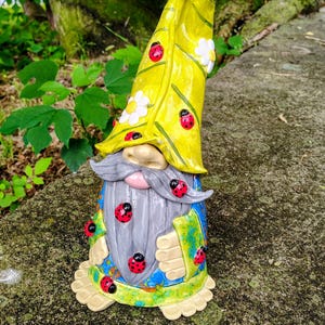 Może przedstawiać: Ceramiczna figurka krasnala ogrodowego z wysoką, spiczastą ż&oacute;łtą czapką ozdobioną białymi kwiatami i czerwonymi biedronkami. Krasnal ma długą szarą brodę, niebieską tunikę z zielonymi akcentami oraz dodatkowe motywy biedronek na brodzie i ubraniu.