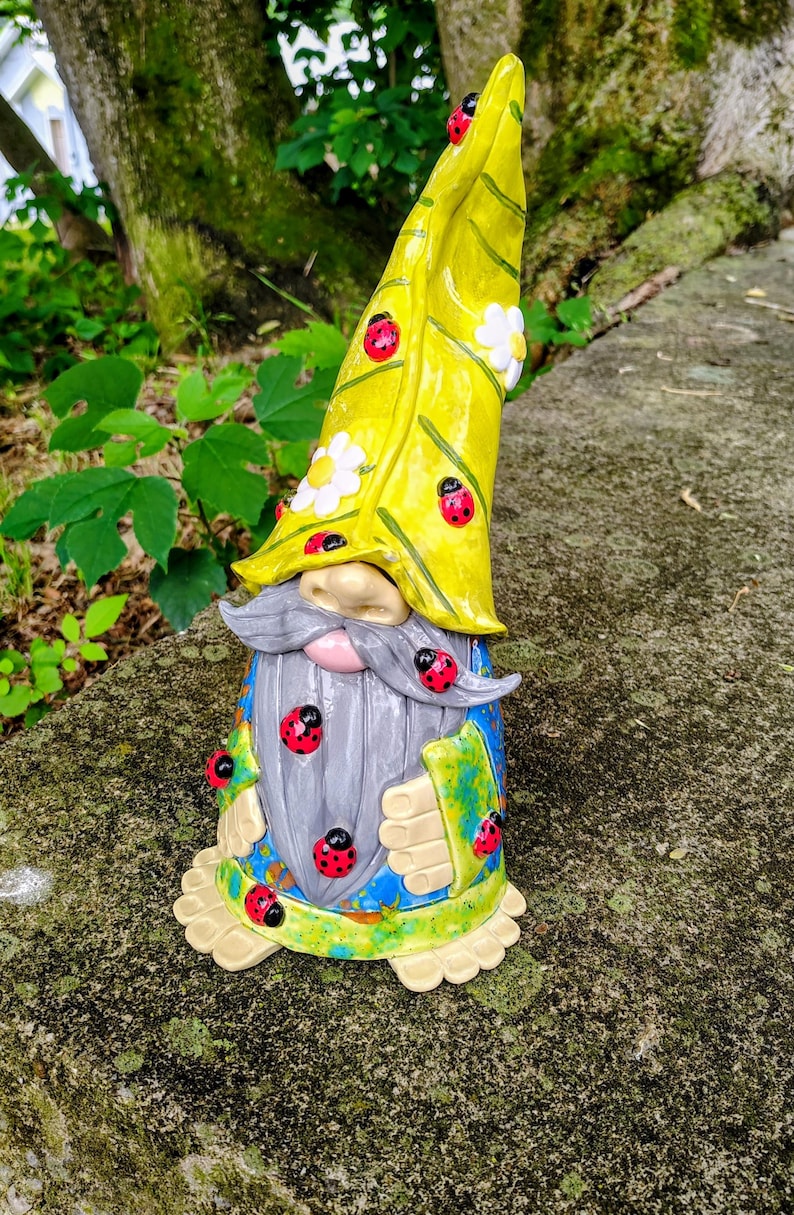 Może przedstawiać: Ceramiczna figurka krasnala ogrodowego z wysoką, spiczastą ż&oacute;łtą czapką ozdobioną białymi kwiatami i czerwonymi biedronkami. Krasnal ma długą szarą brodę, niebieską tunikę z zielonymi akcentami oraz dodatkowe motywy biedronek na brodzie i ubraniu.