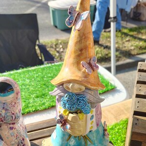 Może przedstawiać: Ceramiczna figurka gnoma z wysokim, spiczastym, marmurkowym pomarańczowym kapeluszem ozdobionym dwoma małymi motylami. Gnom ma szarą brodę, trzyma bukiet niebieskich r&oacute;ż i ma na sobie jasnoniebieskie spodnie z ż&oacute;łtymi akcentami oraz małymi motylami.