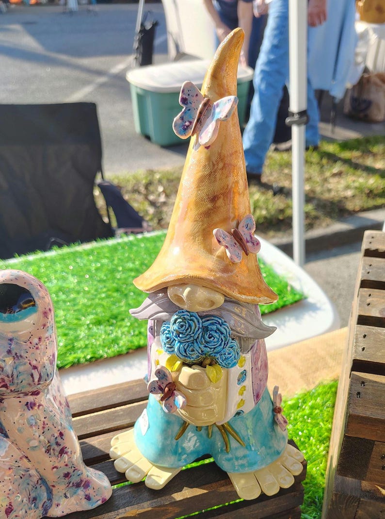 Może przedstawiać: Ceramiczna figurka gnoma z wysokim, spiczastym, marmurkowym pomarańczowym kapeluszem ozdobionym dwoma małymi motylami. Gnom ma szarą brodę, trzyma bukiet niebieskich r&oacute;ż i ma na sobie jasnoniebieskie spodnie z ż&oacute;łtymi akcentami oraz małymi motylami.