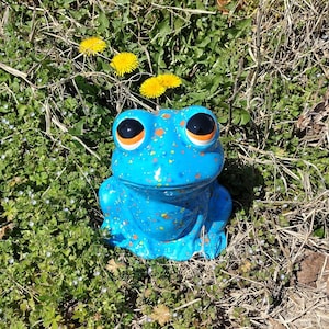 Może przedstawiać: Jasnoniebieska ceramiczna figurka żaby z nakrapianym wzorem w pomarańczowe i żółte kropki. Żaba ma duże, błyszczące czarne oczy z pomarańczowymi obwódkami i siedzi z przednimi łapami opartymi o podłoże.