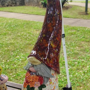 Może przedstawiać: Ceramiczna figurka gnoma z wysoką, spiczastą czapką pokrytą błyszczącym, nakrapianym brązowo-bursztynowym szkliwem. Gnom ma szarą brodę, jasną tunikę w kolorowe kropki oraz jaskrawoniebieskie spodnie. W dłoniach trzyma bukiet pomarańczowych i r&oacute;żowych kwiat&oacute;w.