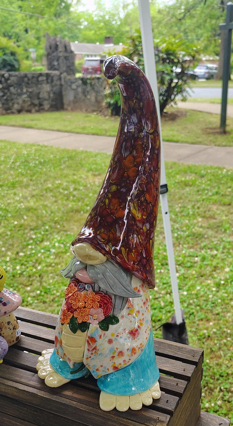 Może przedstawiać: Ceramiczna figurka gnoma z wysoką, spiczastą czapką pokrytą błyszczącym, nakrapianym brązowo-bursztynowym szkliwem. Gnom ma szarą brodę, jasną tunikę w kolorowe kropki oraz jaskrawoniebieskie spodnie. W dłoniach trzyma bukiet pomarańczowych i r&oacute;żowych kwiat&oacute;w.