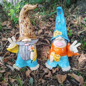 Może przedstawiać: Dwie ceramiczne figurki krasnali w wysokich, spiczastych czapkach. Lewy krasnal ma nakrapianą brązową czapkę, siwą brodę i niebieskie spodnie, trzymając kwiat. Prawy krasnal ma niebieską czapkę w słoneczniki, pomarańczową koszulkę i niebieskie spodnie, trzymając kubek.