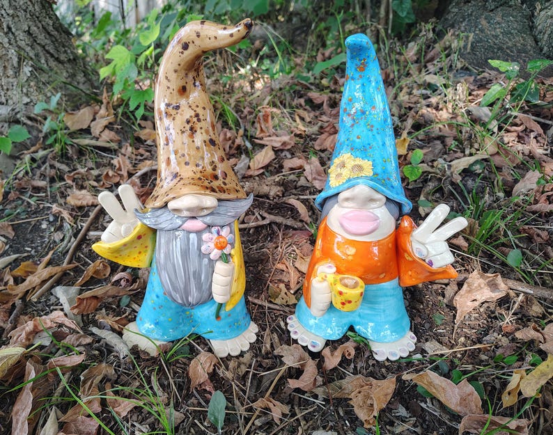 Może przedstawiać: Dwie ceramiczne figurki krasnali w wysokich, spiczastych czapkach. Lewy krasnal ma nakrapianą brązową czapkę, siwą brodę i niebieskie spodnie, trzymając kwiat. Prawy krasnal ma niebieską czapkę w słoneczniki, pomarańczową koszulkę i niebieskie spodnie, trzymając kubek.