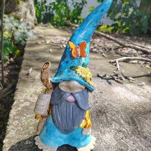 Może przedstawiać: Ceramiczna figurka krasnala ogrodowego z wysoką, niebieską, nakrapianą czapką ozdobioną pomarańczowym motylem i ż&oacute;łtymi słonecznikami. Krasnal ma długą szarą brodę, jasnoniebieską tunikę i trzyma brązową laskę o fakturze przypominającej drewno.