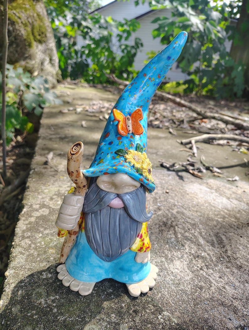 Może przedstawiać: Ceramiczna figurka krasnala ogrodowego z wysoką, niebieską, nakrapianą czapką ozdobioną pomarańczowym motylem i ż&oacute;łtymi słonecznikami. Krasnal ma długą szarą brodę, jasnoniebieską tunikę i trzyma brązową laskę o fakturze przypominającej drewno.
