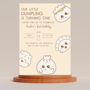 May include: A light beige invitation card with a cartoon dumpling illustration. The text reads "Our little dumpling is turning one. Please join us to celebrate Heather's first birthday. June 15, 2010 at 09 AM. 1234 House Rd, Los Angeles CA. RSVP to Mom 123.456.7890."